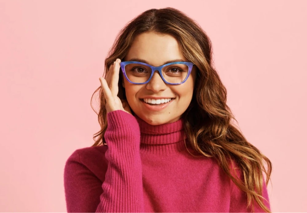 A woman wearing a pink sweater and touching the side of her glasses for heart-shaped faces.