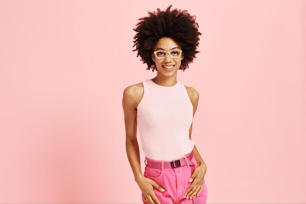 A woman in front of a pink background wearing a pink outfit and rectangular eyeglass frames.